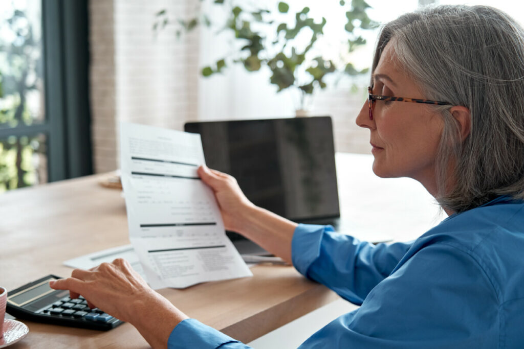 Mature woman at desk looking at document and using calculator.