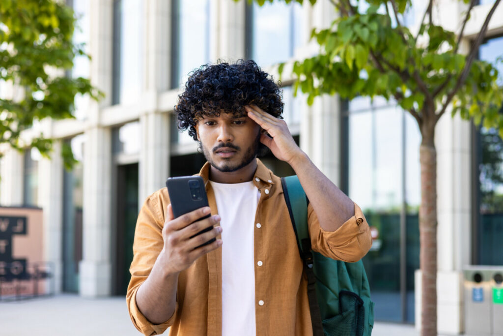 Young college student with a backpack looking anxiously at his smartphone.