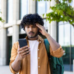 Young college student with a backpack looking anxiously at his smartphone.
