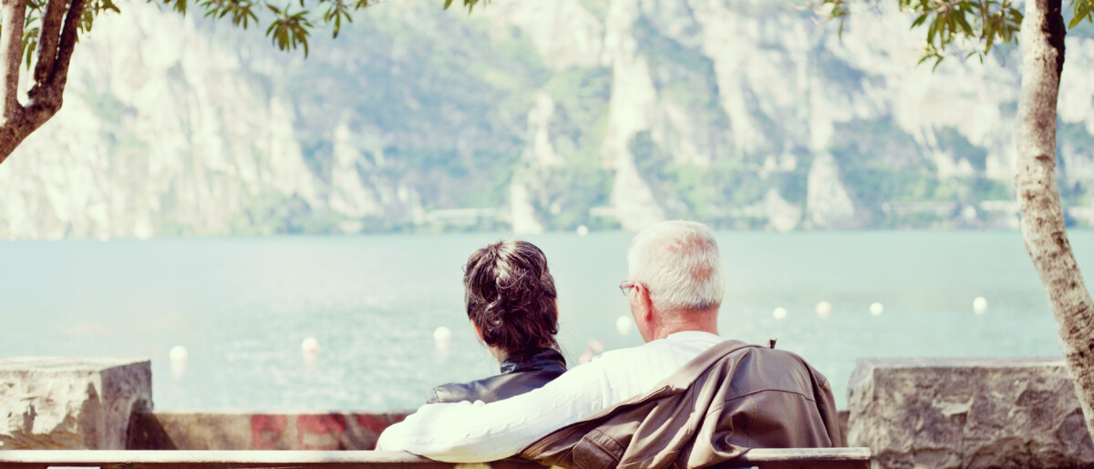 Senior couple sitting and looking out onto the ocean with arms around each other's shoulders.