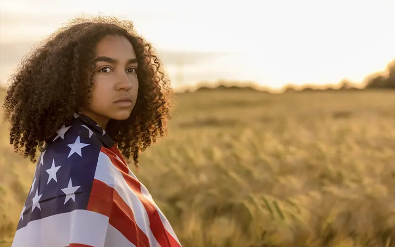 young woman wrapped in an American flag in a field at sunset