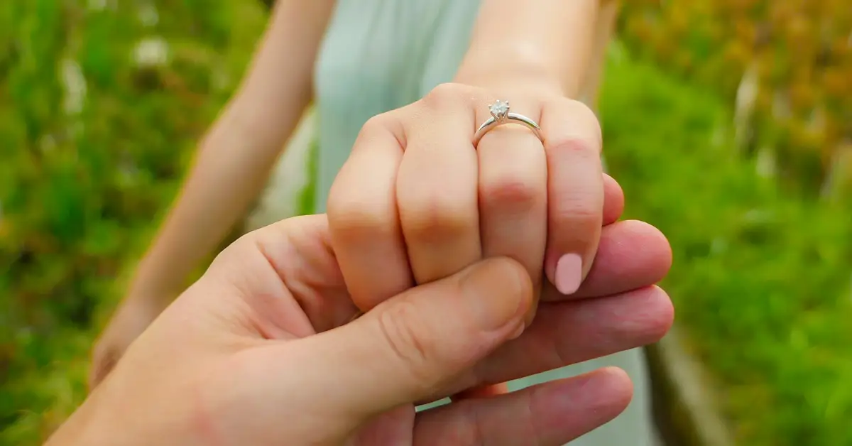 image of a young woman's hand in her fiance's hand in the forefront, showing off her new engagement ring