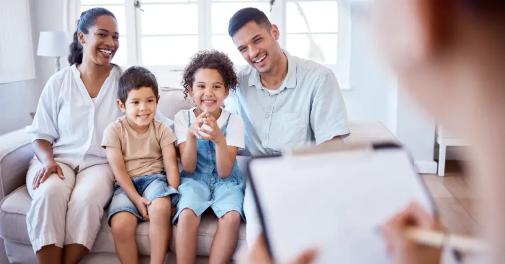 In an office waiting area, a diverse family with children of various ages sits together, likely awaiting their immigration medical examination and discussing vaccination requirements. The scene reflects the importance of vaccination records and moral convictions related to vaccine preventable diseases in the context of immigration services.