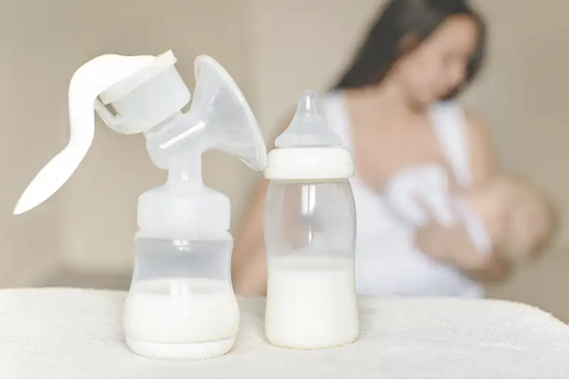 breast pump and breast milk in the foreground with woman breastfeeding in the background