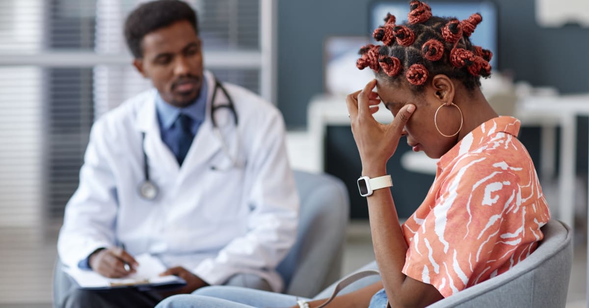 Patient sitting in medical office with head in hand while doctor takes notes, illustrating distress and difficult conversation about a possible medical complication.